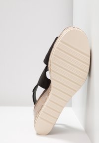 Sandale avec semelle beige rainurée, brides noires et semelle intermédiaire tissée style espadrille, positionnée droite contre le mur.