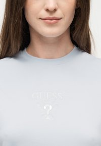 Νέα γυναίκα που φορά μπλε ανοιχτό πουκάμισο με λευκό ανάγλυφο λογότυπο Guess και ερωτηματικό στο ύψος του στήθους, κλειστά χείλη, μακριά καστανά μαλλιά.