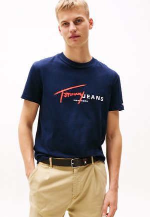 Himmelblaues T-Shirt mit kurzen Ärmeln und einem roten grafischen Logo, auf dem "Tommy Jeans" und "NEW YORK" in Weiß steht. Kombiniert mit khakifarbenen Hosen und einem schwarzen Gürtel.