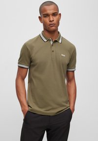 Olivengrønn polo t-skjorte med klassisk krage, to knapper og kontrasterende svart-hvite striper på kragen og ermene. Har logo.