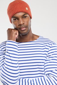 Orange gerippte Strickmütze mit einem Anker-Patch, getragen zu einem weiß-blau gestreiften Langarmshirt. Das Shirt hat einen weiten Halsausschnitt.