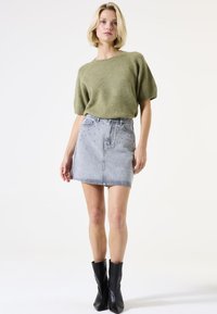 Pull court en tricot vert à manches courtes associé à une jupe en denim gris taille haute ornée de clous argentés. Le mannequin porte des bottines noires.