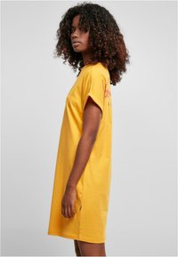 Gul bomulds T-shirt kjole med korte ærmer, løs pasform, sideslids og rød branding på ryggen. Glat tekstur, afslappet design.