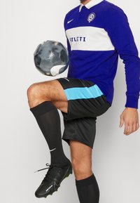Blå långärmad fotbollströja med vit rand, svarta shorts med blå detaljer, svarta strumpor och svarta fotbollsskor som håller i en fotboll.