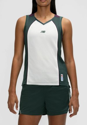 Kvinde iført en ærmeløs hvid og mørkegrøn sportsmesh-top og matchende mørkegrønne mesh-shorts mod en ensfarvet baggrund.