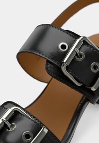 Μαύρο σανδάλι από λουστρίνι με δύο φαρδιές λουρίδες, ρυθμιζόμενες μεταλλικές αγκράφες και καφέ εσωτερική επένδυση. Ομαλή υφή με γυαλιστερό φινίρισμα.