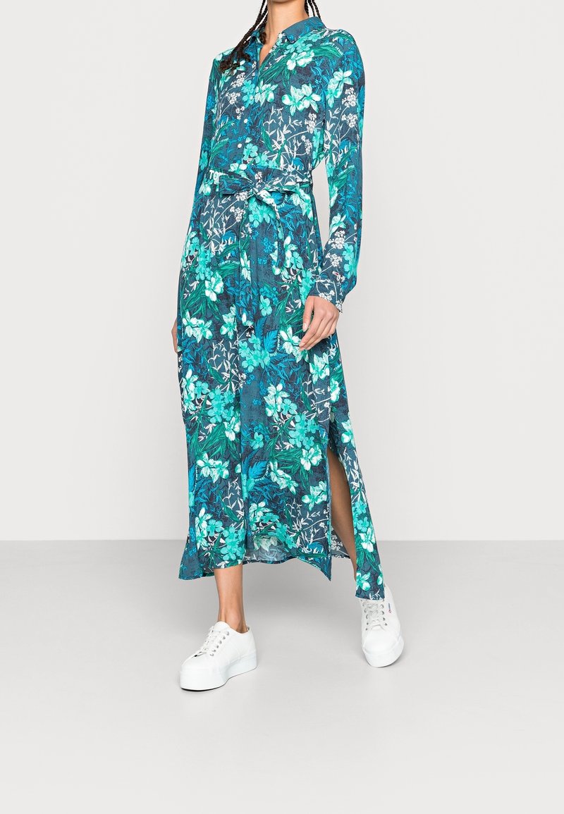Femme portant une robe à manches longues, fleurie bleue et verte, avec une taille nouée et une fente latérale, associée à des baskets blanches à plateforme.