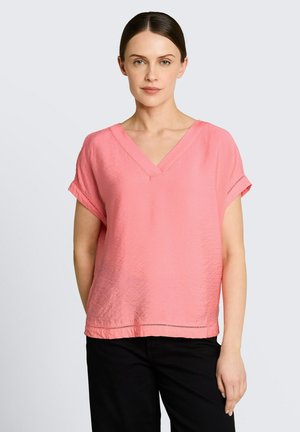 Femme portant un chemisier ample rose à col en V et manches courtes, ainsi qu'un pantalon noir, debout devant un fond gris clair uni.