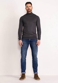 Pulover cu guler înalt, gri închis, cu mâneci lungi, asortat cu blugi albaștri și ghete maro până la gleznă, pozând pe un fundal simplu.