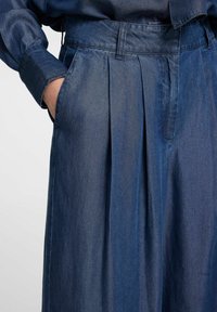 Mörka denimbyxor med veck, som har en mjuk textur, bred midja och sidofickor. Tyget har en subtil glans.
