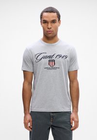 T-shirt cinza de algodão com mangas curtas, apresentando o texto em caligrafia azul-marinho "Gant 1949" e um logotipo em forma de escudo com riscas vermelhas e azuis e estrelas.