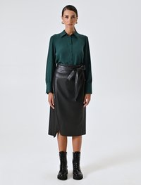 Donna che indossa una camicia a maniche lunghe verde scuro con bottoni, una gonna avvolgente in pelle nera con fiocco, stivali da combattimento neri con lacci e orecchini a cerchio dorati, in piedi dritta.
