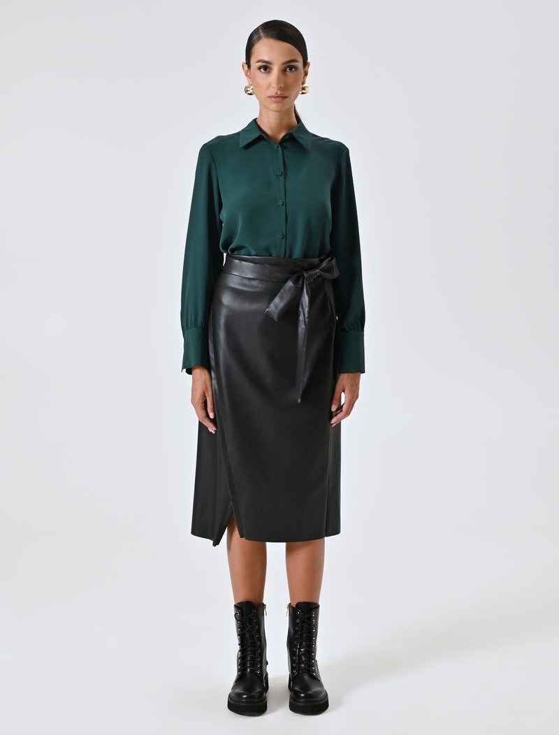 Donna che indossa una camicia a maniche lunghe verde scuro con bottoni, una gonna avvolgente in pelle nera con fiocco, stivali da combattimento neri con lacci e orecchini a cerchio dorati, in piedi dritta.