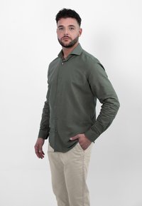 Olijfgroen overhemd met knopen, lange mouwen en een slim fit, gecombineerd met lichtbeige broek. Zachte textuur; subtiele glans.