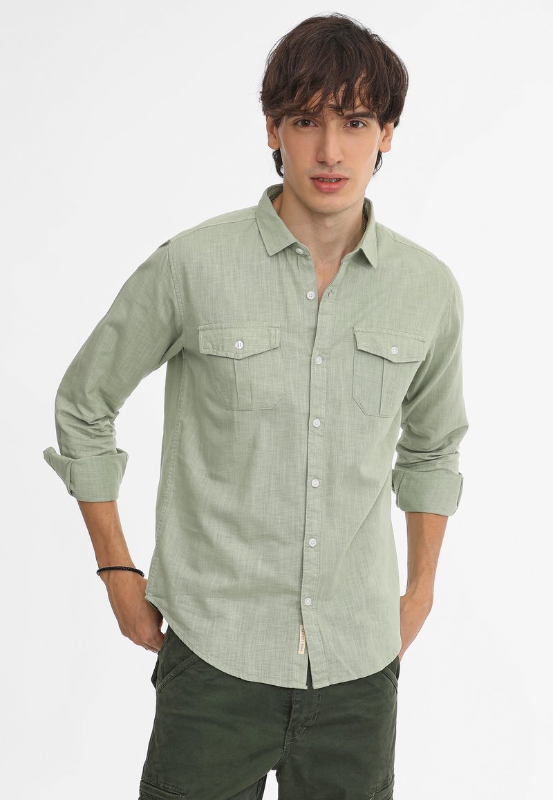 Camisa de botonadura ligera verde hecha de tejido texturizado, que cuenta con dos bolsillos en el pecho, mangas enrollables y botones blancos.