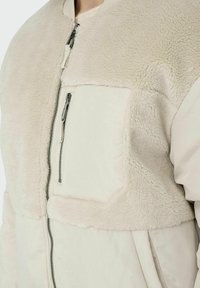 Giacca bomber beige con una texture morbida e pelosa nella parte superiore, dotata di una tasca con zip e contrasto di tessuto tonale.