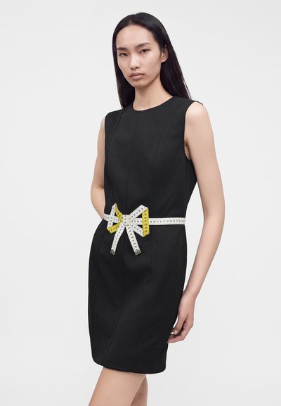 Femme portant une robe noire sans manches avec un ruban à mesurer noué autour de sa taille en forme de nœud sur un fond uni.