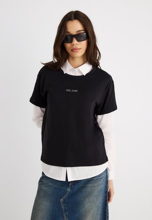 Γυναίκα που φοράει μαύρα γυαλιά ηλίου, μαύρο t-shirt Pepe Jeans πάνω από λευκό πουκάμισο με κουμπιά και μπλε τζιν φούστα, στέκεται μπροστά σε ένα απλό λευκό φόντο.