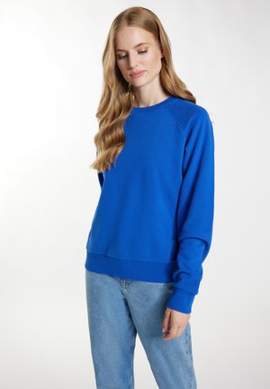 Koninklijk blauwe sweatshirt met lange raglanmouwen, geribbelde manchetten en een aansluitende tailleband. Gecombineerd met lichtblauwe jeans met een klassieke snit.