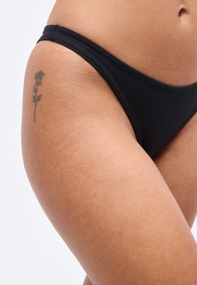 Μαύρο κάτω μέρος μπικίνι από απαλό ύφασμα, με χαμηλό κόψιμο και ελάχιστη κάλυψη, με ένα μικρό τατουάζ τριαντάφυλλου ορατό στο μηρό.