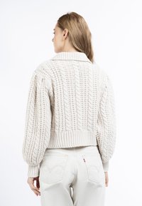 Maglione lavorato a maglia spesso color crema, con colletto, motivi a treccia testurizzati e maniche a palloncino. Vestibilità corta. Indossato con jeans chiari.