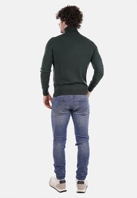 Maglione verde scuro con colletto alto e design aderente. Indossato con jeans azzurri e sneakers bianche, visto da dietro.