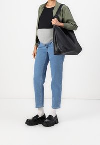 Těhotná žena v neformálním oblečení - modré džíny, černý top, zelená bunda, bílé ponožky, černé robustní mokasíny a velká černá kožená taška.