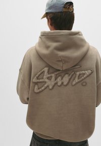 Hanorac bej cu un logo mare, brodat pe spate. Material din amestec de bumbac, cu buzunar tip kangaroo și manșete ribbate.