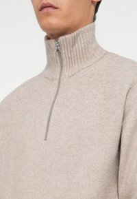 Suéter beige de media cremallera con un cuello alto acanalado. La tela tiene una textura suave y un sutil patrón de tejido, con un detalle de cremallera plateada.