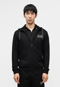 Μαύρο φούτερ με φερμουάρ, κατασκευασμένο από απαλό ύφασμα, με μπροστινή τσέπη, κορδόνια και λογότυπο στο στήθος. Συνδυάζεται με μαύρο παντελόνι που ταιριάζει.