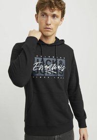 Black hoodie with drawstrings, front pocket, and graphic print featuring "Jack & Jones Essentials" in white and gray. Standard fit.