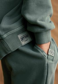 Zelená mikina so rebrovanými manžetami a spodným lemom, s viditeľným označením s textom "Engineered Superdry Heavy Duty." Mäkká textúra tkaniny.
