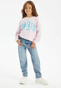 Svetlo roza pulover s plavim tiskom "PARIS", v kombinaciji z svetlo modrimi ohlapnimi kavbojkami in belimi športnimi copati. Mehak material, sproščen kroj.