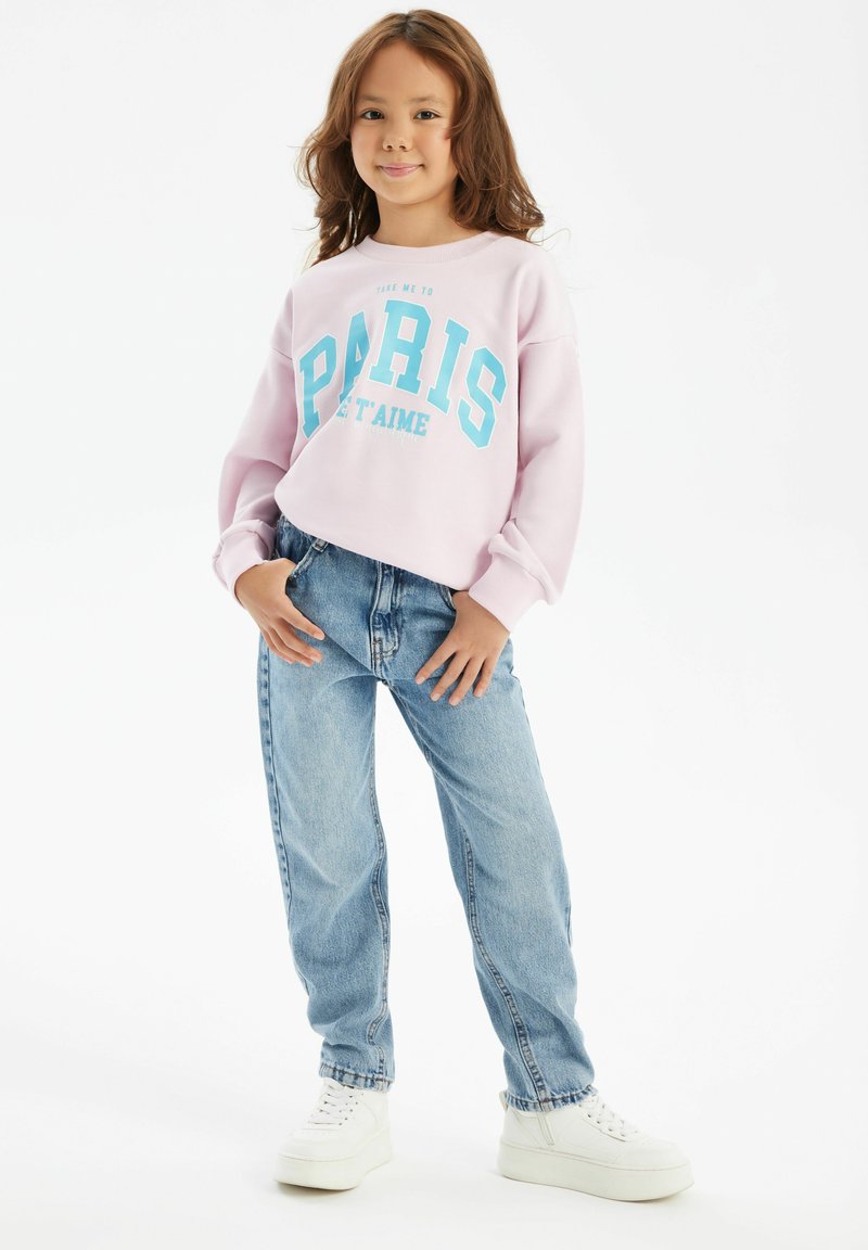 Svetlo roza pulover s plavim tiskom "PARIS", v kombinaciji z svetlo modrimi ohlapnimi kavbojkami in belimi športnimi copati. Mehak material, sproščen kroj.