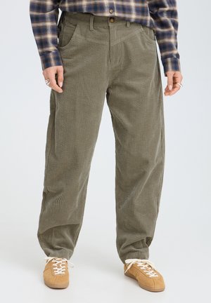 Persona che indossa pantaloni di velluto a coste verdi, sneakers beige con lacci bianchi e una camicia a quadri blu e marrone, in piedi contro uno sfondo uniforme.