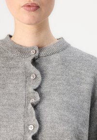 Grå cardigan med en bølget knappestolpe, med runde plastknapper. Myk tekstur og ribbet halslinning. Enkelt design.