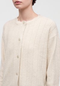 Femme portant un cardigan tricoté couleur crème avec des rayures verticales texturées et des boutons ronds, montrée des épaules jusqu’au milieu du torse.