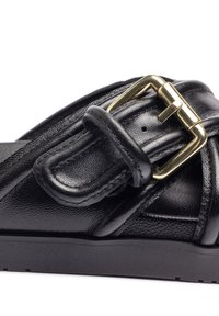 Zwarte leren sandaal met een kruisontwerp, voorzien van een gouden gesp en een gestructureerd oppervlak. De zool is vlak en gemaakt van rubber.