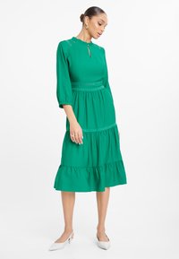 Abito midi verde con orlo a volant, rifiniture in pizzo e scollatura a chiave. Maniche a tre quarti e silhouette rilassata. Décolleté bianche.