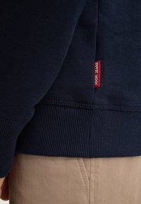 Felpa blu navy con texture a coste all'orlo e ai polsini, presenta un'etichetta rossa "JOOP! JEANS" sul lato. Abbinata a tessuto beige.