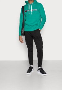 Sinivihreä hupullinen collegepaita valkoisilla nyöreillä ja logolla; mustat kapeat vetoketjuhousut taskuilla; mustat lenkkarit valkoisilla yksityiskohdilla.