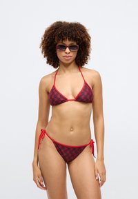 Raudonas ir juodas bikinis su trikampiu viršumi ir raišteliais šonuose. Yra kartojama logotipo raštas ir plonos raudonos dirželiai. Lygus audinio paviršius.