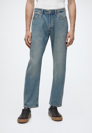 Personne portant un jean droit bleu clair, des chaussettes blanches et des baskets noires avec des semelles marron, debout sur un fond blanc uni.