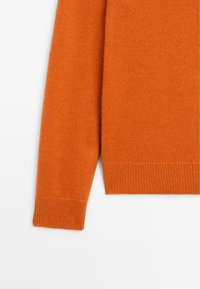Manche de pull tricoté orange avec bord-côte aux poignets et à l'ourlet sur fond blanc.