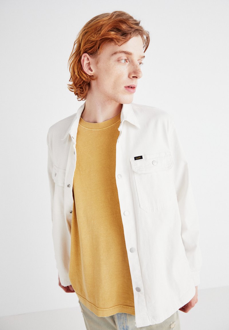 Camisa blanca de botones con un bolsillo, superpuesta a una camiseta de color mostaza. Jeans de mezclilla azul claro desgastados, sin patrones ni acentos visibles.