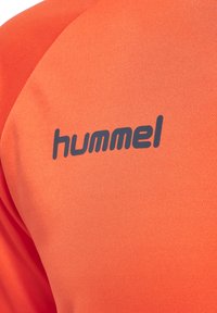Maglietta sportiva arancione con tessuto testurizzato e finitura liscia, caratterizzata da un logo "hummel" blu scuro stampato sulla spalla.