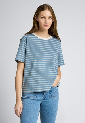 Junge Frau mit hellbraunen Haaren, die ein blau-weiß gestreiftes Kurzarmhemd und blaue Jeans trägt, eine Hand in der Tasche, neutraler Hintergrund.