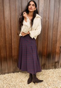 Krém színű, kötött mintás pulóver széles gallérral, hosszú bordó kockás szoknyával és barna hegyes orrú csizmával párosítva. Fa hátterű.