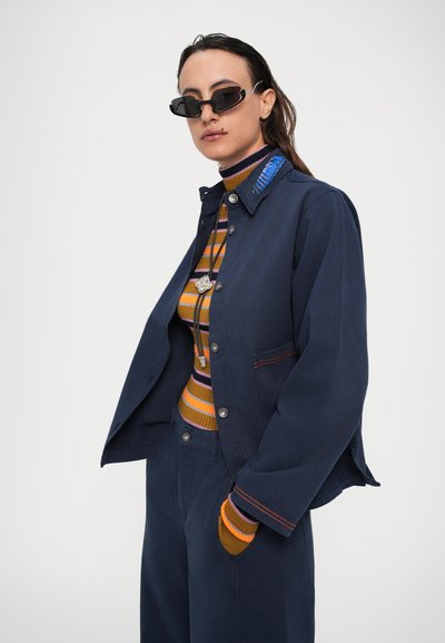 Άτομο που φοράει σκούρο μπλε σακάκι και παντελόνι, ριγέ ζακέτα με τσέπη, γυαλιά ηλίου και μακρύ κολιέ, μπροστά σε λευκό φόντο.