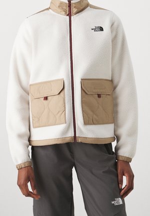 Osoba ubrana w biały polarowy sweter z jasnobrązowymi, pikowanymi kieszeniami i kołnierzem, w połączeniu z ciemnoszarymi spodniami, obie marki The North Face.